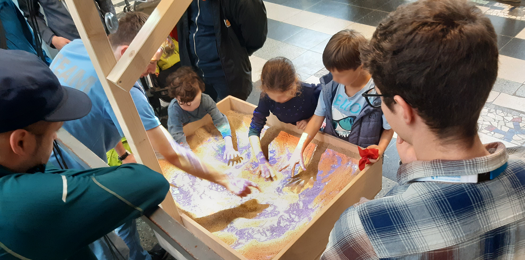 drei Kinder zeichnen Landschaften in einer Sandbox die mittels Licht Landschaftsveränderungen simulieren kann, ein Erwachsener erklärt und mehrere weitere Erwachsene beobachten
